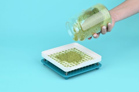 Hand pouring herbal powder into a manual capsule filling machine on a blue background.